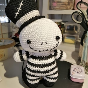 May include: A crocheted skeleton doll wearing a black and white striped outfit and a black top hat with white stitching. The doll has black eyes and a stitched-on smile.