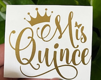 May include: Gold vinyl decal with the text "Mis Quince" in a cursive font with a crown above the text and a butterfly to the right of the text.