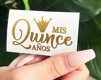 May include: A white rectangular sticker with gold lettering and a crown design. The text reads "Mis Quince Años". The sticker is held against a background of green foliage.