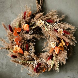 Könnte beinhalten: Ein kreisförmiger Kranz aus getrocknetem Pampasgras, Weizenhalmen und kleinen weißen Blumen. Das Arrangement enthält Akzente aus dunkelrotem, leuchtend orangefarbenem und goldgelbem Trockenlaub und ist an einer dicken, gedrehten Juteschnur aufgehängt.