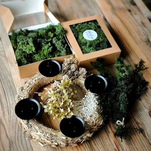 Könnte beinhalten: Ein Strohkranz, verziert mit getrockneten beigen Blumen und vier schwarzen runden Kerzenhaltern. Daneben stehen zwei Pappschachteln mit grünem konserviertem Moos, eine offen und eine geschlossen mit Sichtfenster und rundem Etikett.