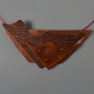 Pode incluir: Um colar de cobre com um design geométrico. O pingente é feito de três peças de cobre sobrepostas, cada uma com uma superfície texturizada. O colar é enfiado em um cordão rosa.