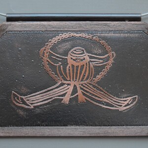 Puede incluir: Un collar con un colgante de cobre, adornado con un dije ovalado de color marrón oscuro que representa una figura estilizada con un diseño circular alrededor de la cabeza. La figura está grabada en cobre sobre el fondo marrón oscuro.