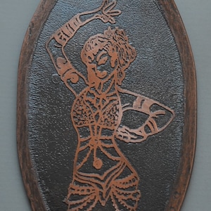 Puede incluir: Un colgante de cobre con un marco de madera marrón oscuro. El colgante presenta un grabado detallado de una mujer en una pose de baile.