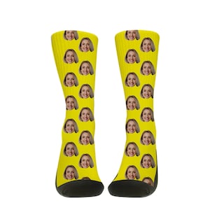 May include: A pair of yellow socks with a repeating pattern of a smiling woman's face. The socks have black heels and toes. The socks are made of a soft, comfortable material.