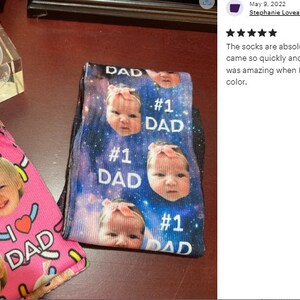 Pu&ograve; includere: Due paia di calzini con disegni personalizzati di facce di bambino. Un paio &egrave; rosa con testo "I LOVE DAD" e facce di bambino. L'altro paio &egrave; nero con sfondo galassia, facce di bambino e testo "#1 DAD".