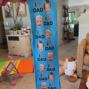 Pu&ograve; includere: Un paio di calzini blu con la scritta "I ❤️ DAD" e molteplici immagini di volti di bambini. I calzini sono tenuti davanti a uno sfondo sfocato.