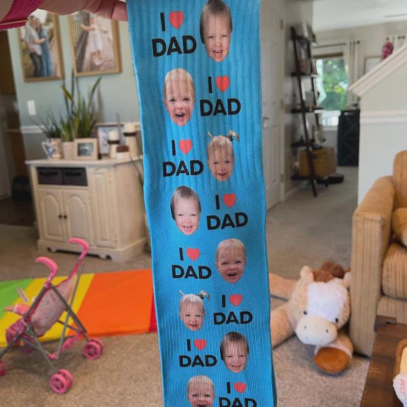 Pu&ograve; includere: Un paio di calzini blu con la scritta "I ❤️ DAD" e molteplici immagini di volti di bambini. I calzini sono tenuti davanti a uno sfondo sfocato.