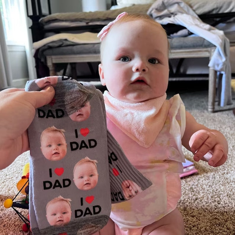 Pu&ograve; includere: Un bambino con un bavaglino e un body rosa accanto a un paio di calzini grigi. I calzini hanno le parole "DAD" e "I ❤️ DAD" stampate, insieme alle immagini del viso del bambino.