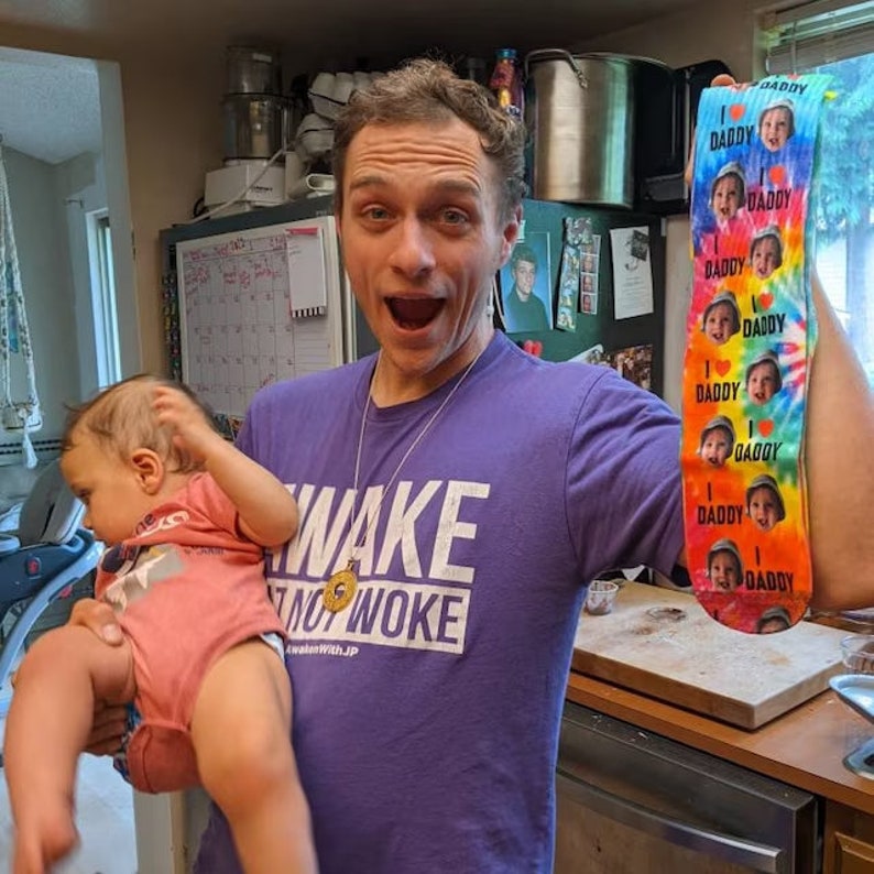 Pu&ograve; includere: Un paio di calzini colorati con un motivo tie-dye e le parole "I ❤️ DADDY" ripetute, insieme al volto di un bambino. I calzini sono tenuti da una persona che indossa una maglietta viola con le parole "AWAKE NOT WOKE".