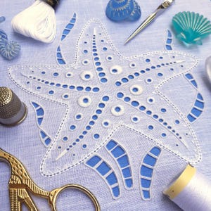 May include: A white embroidered starfish design on a light blue background. The starfish has a detailed, openwork pattern with small, embroidered circles. Sewing tools and decorative sea-themed items, such as a thimble and scissors, are also visible.