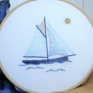 May include: Embroidered sailboat design in a wooden hoop. The sailboat features blue sails with detailed stitching, a gold-colored accent, and is set against a white background. A stitched sun is in the upper right corner. The hoop measures approximately 8 inches in diameter.