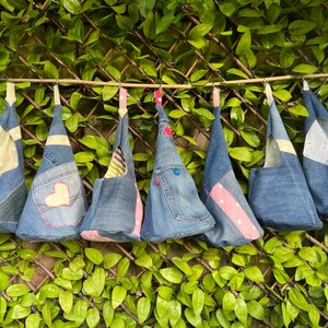 May include: Seven denim bags hanging from a wooden dowel. The bags are made from repurposed denim and have different fabric accents, including hearts, polka dots, and floral patterns.