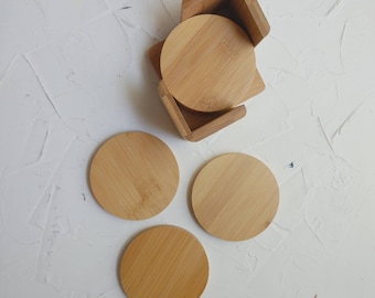 May include: Set of four round bamboo coasters with a wooden holder. The coasters are light brown and have a natural wood grain.