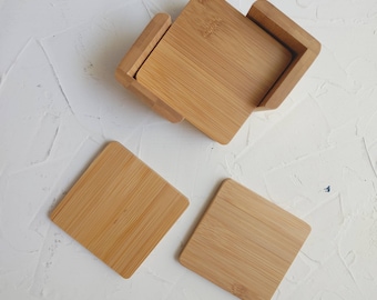 May include: Set of four square bamboo coasters. The coasters are stacked in a square shape, with three coasters visible. The coasters are a light brown colour and have a smooth, natural finish.