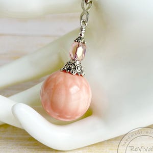 May include: A pink gemstone pendant necklace with a silver chain. The pendant features a round, textured pink stone, a silver filigree cap, and a pink crystal bead. The necklace is displayed on a white hand.