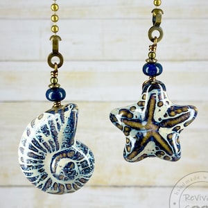May include: Two ceramic fan pull chains, one shaped like a seashell and the other like a starfish. Both are white with blue and brown accents. The chains are gold and have blue beads.
