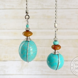 May include: Two turquoise and brown beaded light pull chains with a silver chain and a silver hook at the top. The beads are round and have a crackled finish. The chains are hanging against a white wood background.