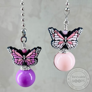 May include: Two butterfly-themed ceiling fan pull chains. Each features a butterfly with pink and white wings and a black outline, atop a round, colored ball. One ball is purple, the other is pink. Both are suspended by a silver chain.