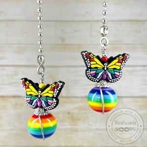 May include: Two butterfly-themed light pull chains. Each chain features a colorful butterfly charm with yellow, red, and black accents, atop a rainbow-striped sphere. The chains are suspended from a silver ball chain.