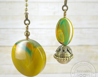 May include: Two decorative ceiling fan pull chains. Each chain has a brass-colored metal chain and a decorative pull. The pulls are oval-shaped with yellow, green, and white swirls. One pull has a decorative metal base.