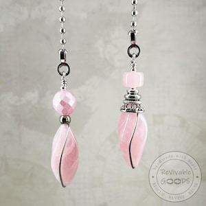 May include: Two ceiling fan pulls featuring light pink beads and teardrop-shaped pendants. Each pull hangs from a silver-toned metal ball chain. The left pull has a faceted round bead, while the right pull features a smooth square bead and a decorative silver cap above the pink teardrop. A circular logo with the text Revivable GOODS is visible in the bottom right corner.