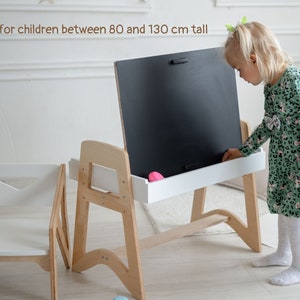 May include: A wooden easel with a chalkboard and a white shelf. A child is standing at the easel, reaching for a pink eraser. The easel is designed for children between 80 and 130 cm tall.