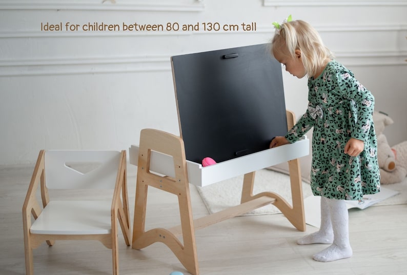 May include: A wooden easel with a chalkboard and a white shelf. A child is standing at the easel, reaching for a pink eraser. The easel is designed for children between 80 and 130 cm tall.