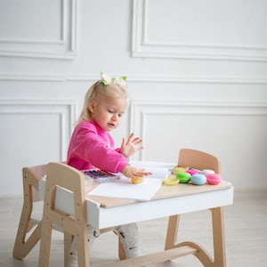 May include: A young girl sits at a white and wood children's desk and chair set. She is wearing a pink sweater and is playing with play dough. There are colorful crayons and macarons on the desk.