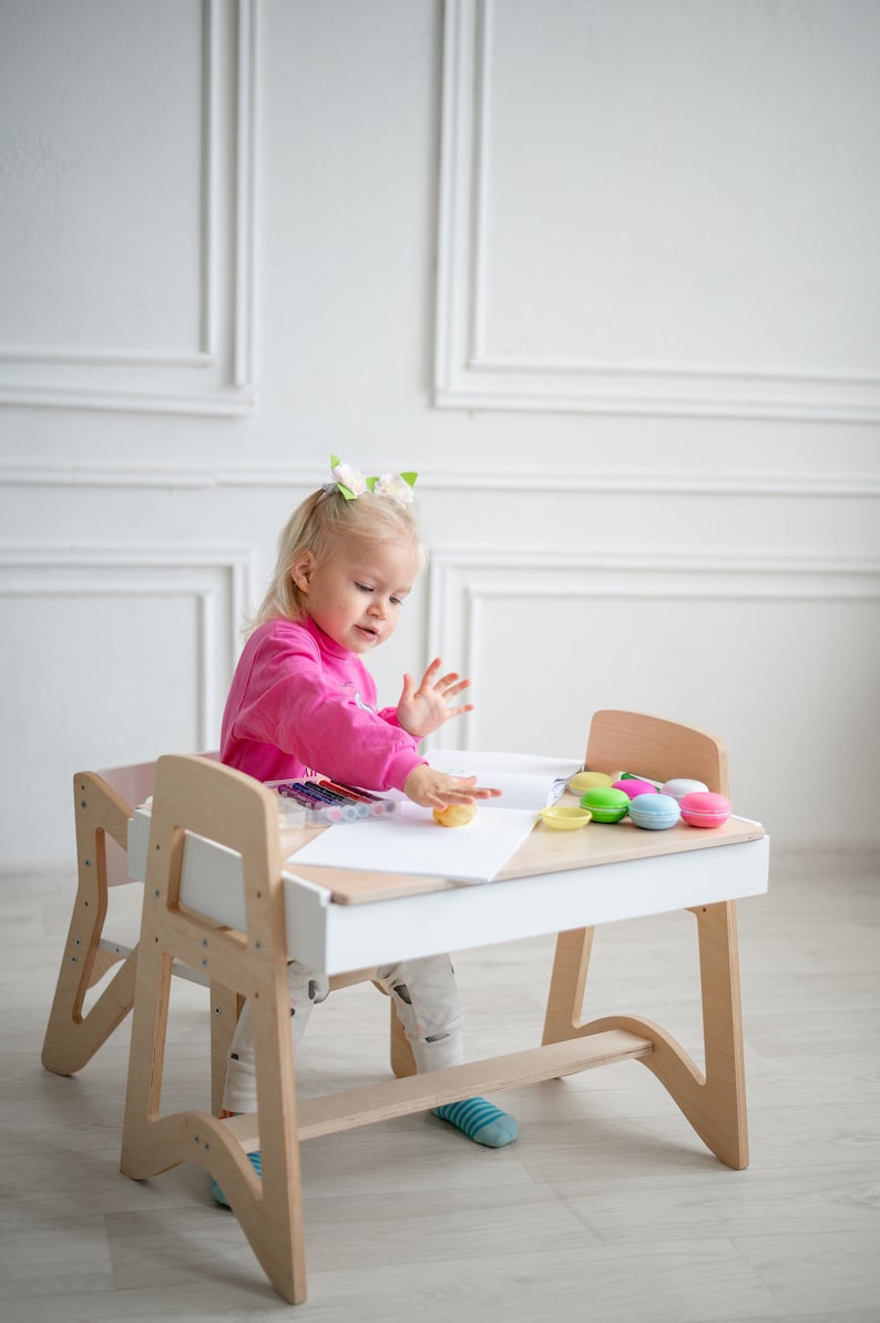 May include: A young girl sits at a white and wood children's desk and chair set. She is wearing a pink sweater and is playing with play dough. There are colorful crayons and macarons on the desk.