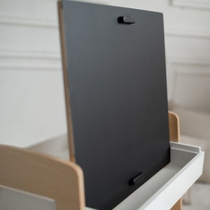 May include: A wooden easel with a black chalkboard surface and a white drawer. The chalkboard is hinged and can be flipped up to reveal the drawer.