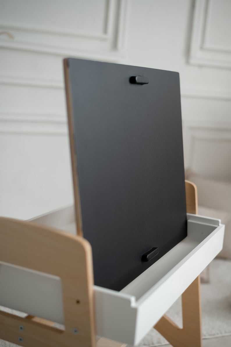 May include: A wooden easel with a black chalkboard surface and a white drawer. The chalkboard is hinged and can be flipped up to reveal the drawer.