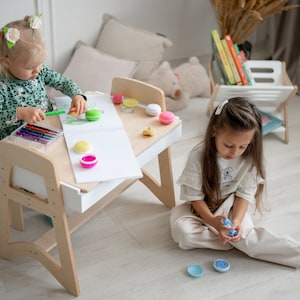 May include: Two children playing at a wooden table with a white top. The table has a built-in drawer and a small chair. The girl on the left is drawing with crayons on a white sheet of paper. The girl on the right is playing with colorful plastic toys.