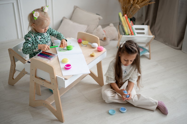 May include: Two children playing at a wooden table with a white top. The table has a built-in drawer and a small chair. The girl on the left is drawing with crayons on a white sheet of paper. The girl on the right is playing with colorful plastic toys.