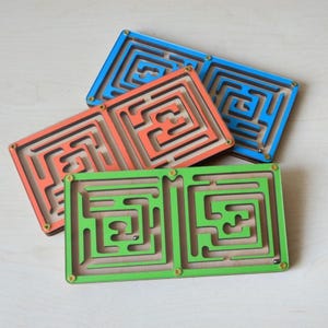 May include: Three wooden maze games with different colored borders. The mazes are made of wood and have a groove for a marble to roll through. The mazes are stacked on top of each other, with the green maze on the bottom, the orange maze in the middle, and the blue maze on top.