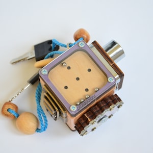 Puede incluir: Un rompecabezas de madera con una ventana de plástico transparente y una perilla de metal. El rompecabezas tiene una serie de pequeñas bolas de metal que se pueden mover dentro de la ventana. El rompecabezas está unido a un cordón azul con cuentas de madera.