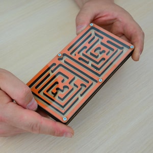 May include: A wooden maze puzzle with two levels, each with a complex orange and black design. The maze is held in the hands of a person.