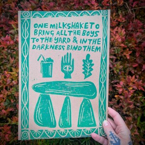 K&ouml;nnte beinhalten: Gr&uuml;ner und brauner Druck mit dem Text "ONE MILKSHAKETO BRING ALL THE BOYS TO THE YARD & IN THE DARKNESS BIND THEM". Der Druck zeigt einen Milchshake, eine Hand mit einem Auge, ein Eichenblatt und ein stilisiertes Bild einer Steinstruktur.