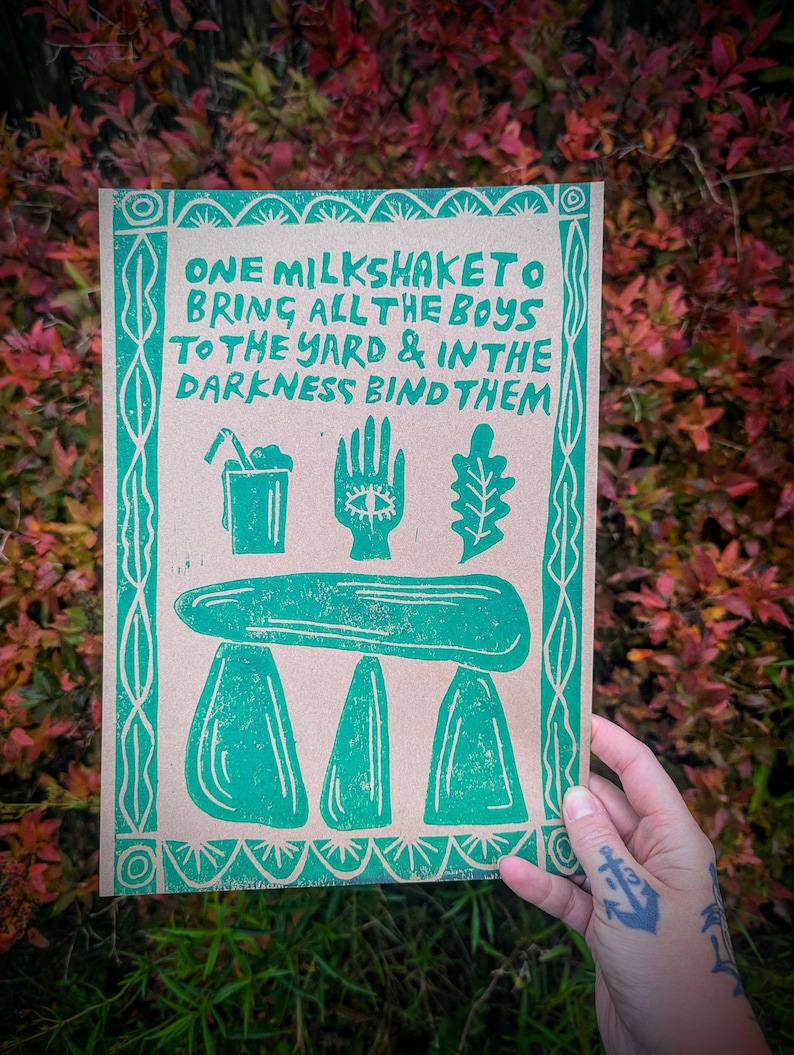 K&ouml;nnte beinhalten: Gr&uuml;ner und brauner Druck mit dem Text "ONE MILKSHAKETO BRING ALL THE BOYS TO THE YARD & IN THE DARKNESS BIND THEM". Der Druck zeigt einen Milchshake, eine Hand mit einem Auge, ein Eichenblatt und ein stilisiertes Bild einer Steinstruktur.