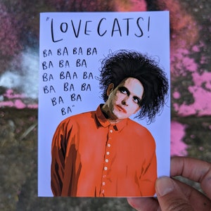 May include: A white card with the text "LOVE CATS!" and a black and white illustration of a person with black hair wearing a bright orange button-down shirt. The text "BA BA BA BA BA BA BA BAA BA BA BA BA BA" is printed below the illustration.