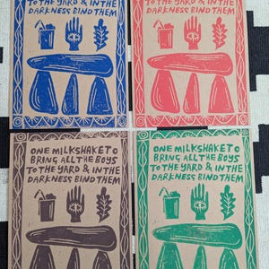 K&ouml;nnte beinhalten: Vier braune Papierkarten mit wei&szlig;em Rand. Jede Karte hat ein anderes farbiges Design mit dem Text "ONE MILKSHAKETO BRING ALL THE BOYS TO THE YARD & IN THE DARKNESS BIND THEM". Die Designs sind blau, rot, braun und gr&uuml;n.