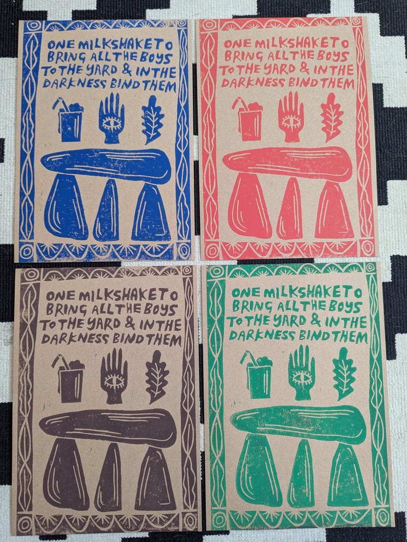 K&ouml;nnte beinhalten: Vier braune Papierkarten mit wei&szlig;em Rand. Jede Karte hat ein anderes farbiges Design mit dem Text "ONE MILKSHAKETO BRING ALL THE BOYS TO THE YARD & IN THE DARKNESS BIND THEM". Die Designs sind blau, rot, braun und gr&uuml;n.