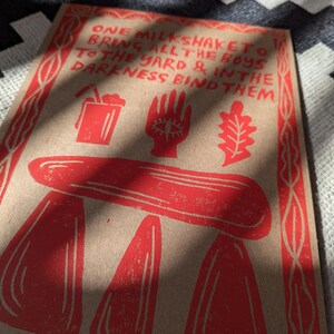 K&ouml;nnte beinhalten: Rote und wei&szlig;e Illustration auf braunem Hintergrund mit dem Text "ONE MILKSHAKETO BRING ALL THE BOYS TO THE YARD & IN THE DARKNESS BIND THEM".