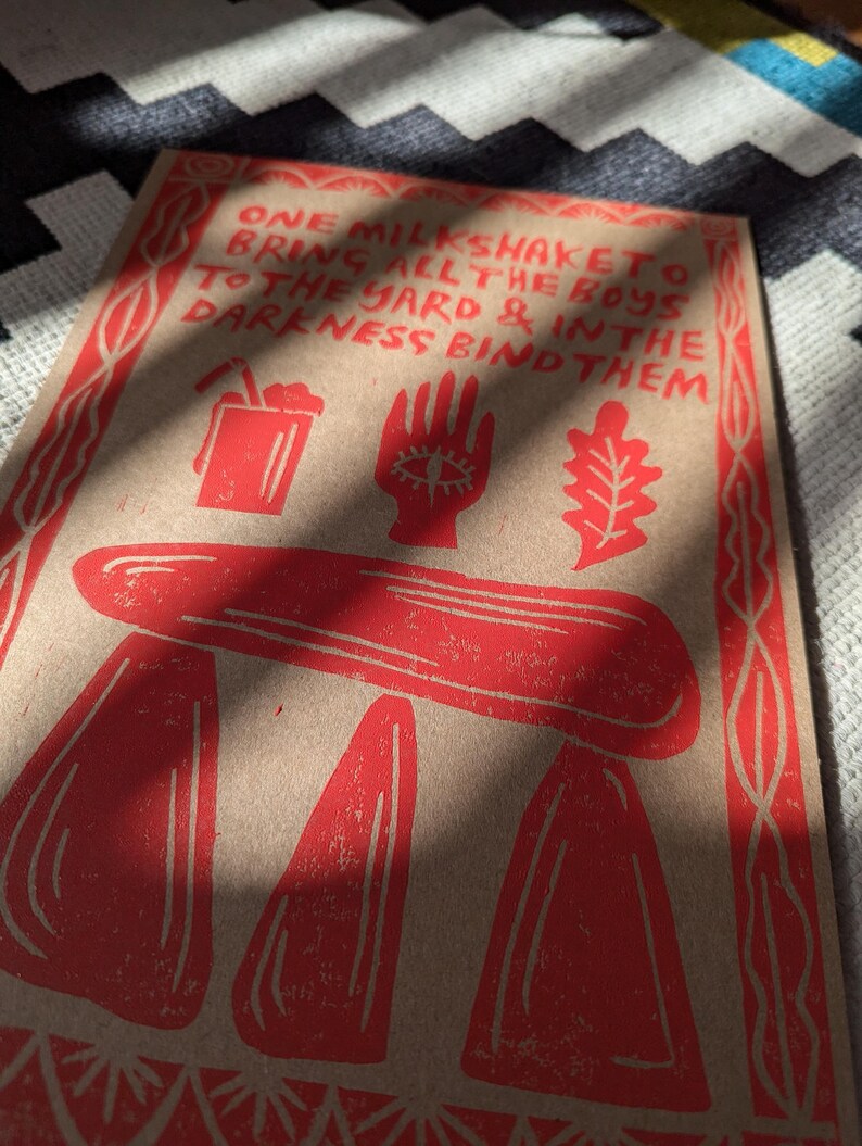 K&ouml;nnte beinhalten: Rote und wei&szlig;e Illustration auf braunem Hintergrund mit dem Text "ONE MILKSHAKETO BRING ALL THE BOYS TO THE YARD & IN THE DARKNESS BIND THEM".