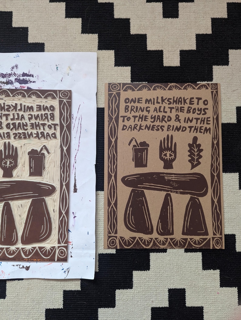 K&ouml;nnte beinhalten: Ein brauner Papierdruck mit dem Text "ONE MILKSHAKE TO BRING ALL THE BOYS TO THE YARD & IN THE DARKNESS BIND THEM". Der Druck zeigt eine Hand mit einem Auge, eine Tasse mit einem Strohhalm, ein Blatt und eine dreisteinige Struktur.