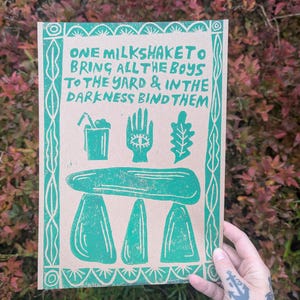 K&ouml;nnte beinhalten: Gr&uuml;ner und wei&szlig;er Druck auf braunem Papier mit dem Text "ONE MILKSHAKETO BRING ALL THE BOYS TO THE YARD & IN THE DARKNESS BIND THEM". Der Druck zeigt eine Hand mit einem Auge, ein Blatt, ein Glas mit einem Strohhalm und einen dreibeinigen Hocker.