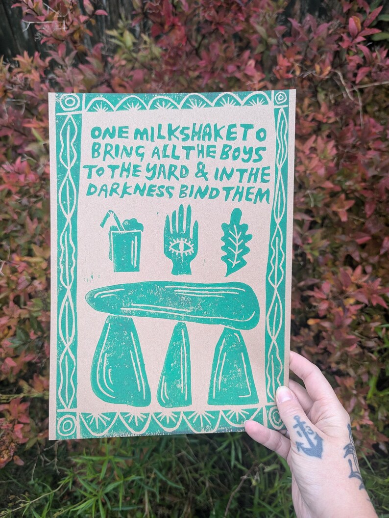 K&ouml;nnte beinhalten: Gr&uuml;ner und wei&szlig;er Druck auf braunem Papier mit dem Text "ONE MILKSHAKETO BRING ALL THE BOYS TO THE YARD & IN THE DARKNESS BIND THEM". Der Druck zeigt eine Hand mit einem Auge, ein Blatt, ein Glas mit einem Strohhalm und einen dreibeinigen Hocker.