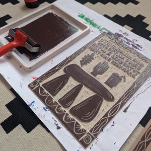 K&ouml;nnte beinhalten: Ein brauner Linolschnitt mit einem Design aus drei Pilzen, einem Tisch, einer Hand, einem Blatt und einem Eimer. Der Druck befindet sich auf einem wei&szlig;en Blatt Papier mit einem braunen Walzenroller und einem roten Stempelkissen im Hintergrund.