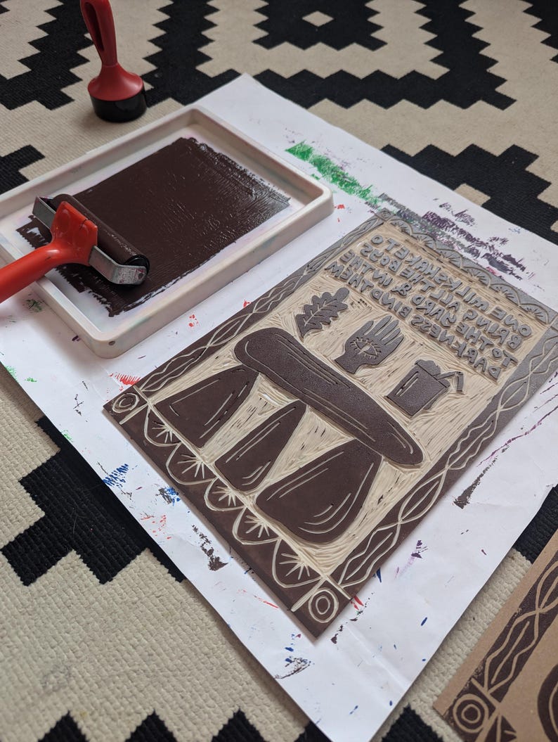 K&ouml;nnte beinhalten: Ein brauner Linolschnitt mit einem Design aus drei Pilzen, einem Tisch, einer Hand, einem Blatt und einem Eimer. Der Druck befindet sich auf einem wei&szlig;en Blatt Papier mit einem braunen Walzenroller und einem roten Stempelkissen im Hintergrund.
