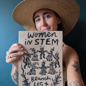 Peut inclure: Une personne portant un chapeau de paille tient une estampe en papier brun avec de l'encre noire. L'estampe représente un cercle de figures en noir, avec le texte "Women in STEM" en haut et "Branch Log + Bog" en bas.